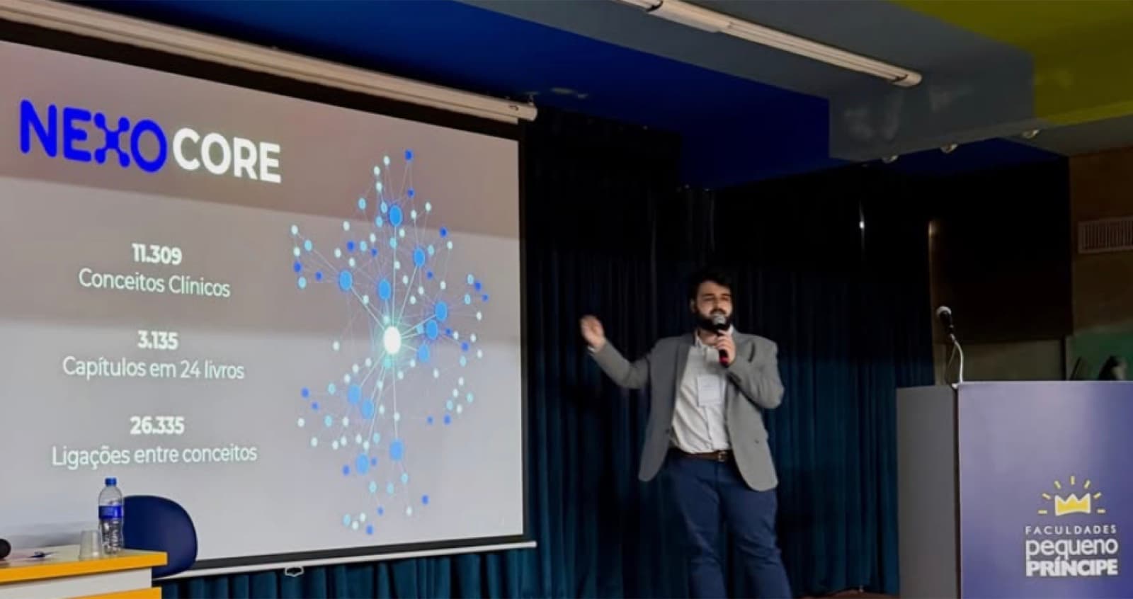 Thiago Moraes al micrófono frente a una pantalla con la diapositiva de NEXO Core y un grafo de conceptos clínicos, en el auditorio de las Faculdades Pequeno Príncipe.