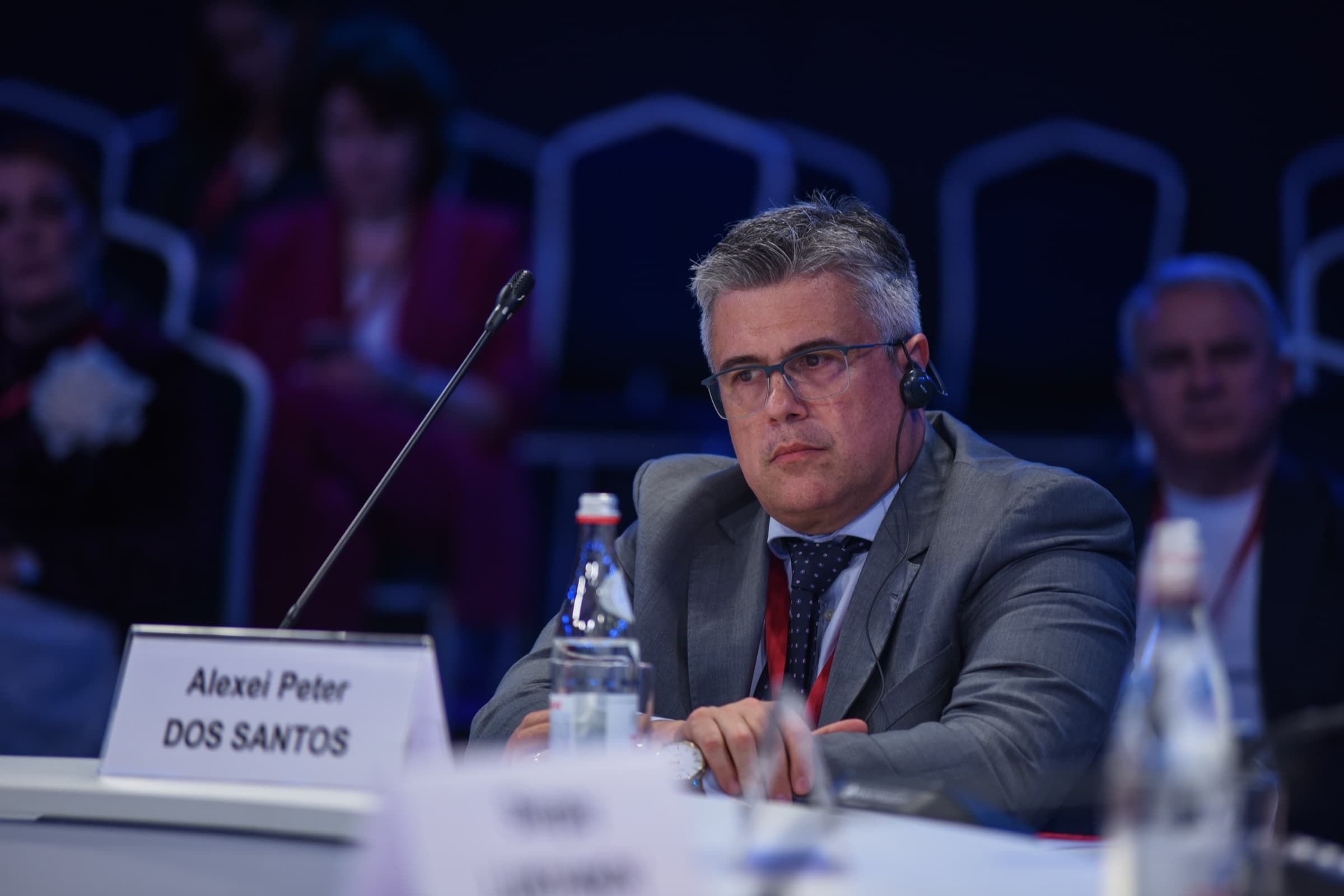 El Dr. Alexei Peter sentado a la mesa de la sesión, con auricular de traducción simultánea y su placa de identificación delante.