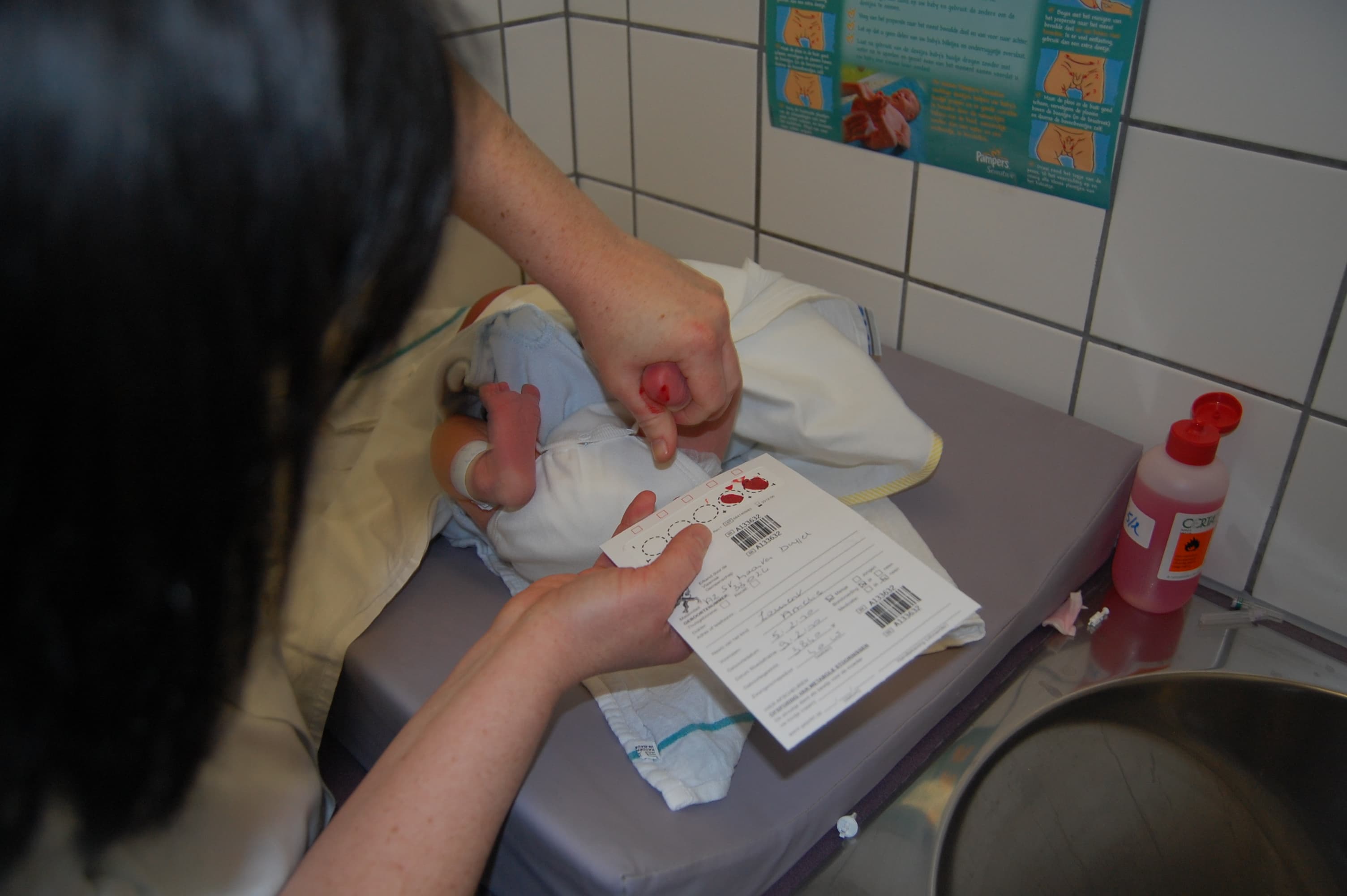 Prueba del talón en Flandes, Bélgica: una mano sostiene el pie del recién nacido, con la gota de sangre en el talón, y la otra sostiene la tarjeta de papel filtro mientras se llenan los círculos.