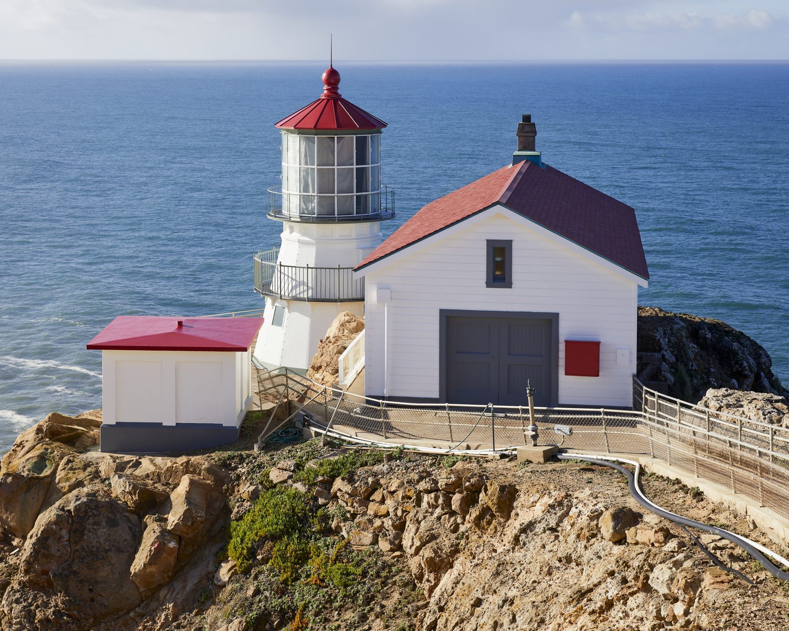 Farol branco de Point Reyes, na Califórnia, com telhado vermelho, sobre um rochedo à beira do mar azul num dia claro.