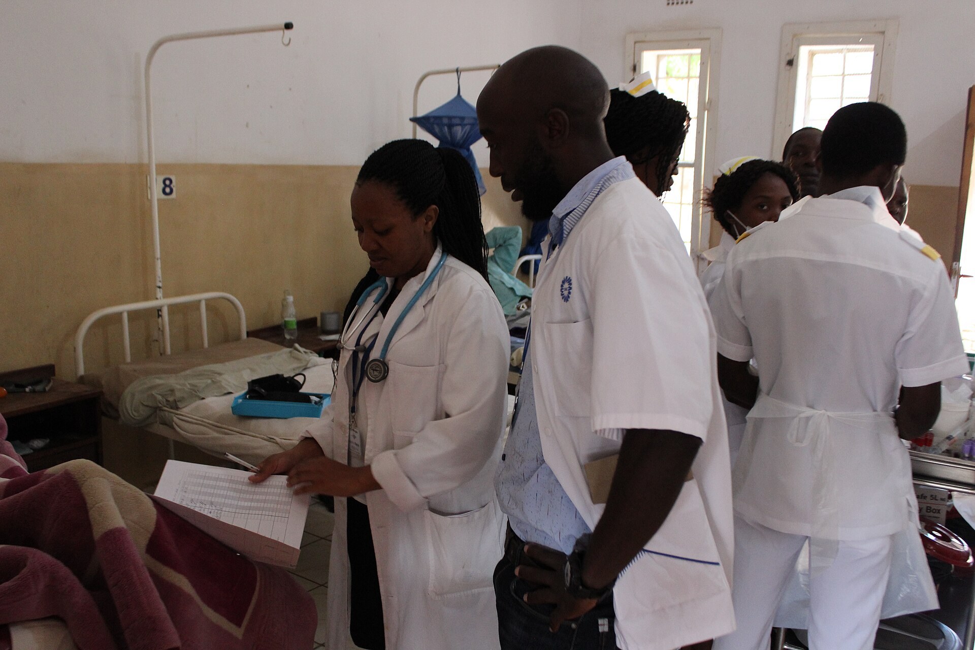 A doctor in a white coat with a blue stethoscope reads a sheet of notes beside a bed, while a colleague in a white coat and nurses in white uniforms look on, in a ward with light-colored walls.