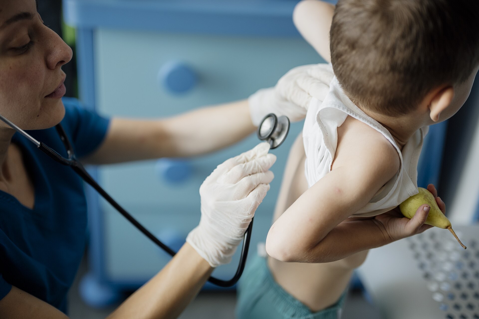 Profesional de salud con guantes y uniforme azul ausculta la espalda de un niño que sostiene una pera, en una sala de consultorio.