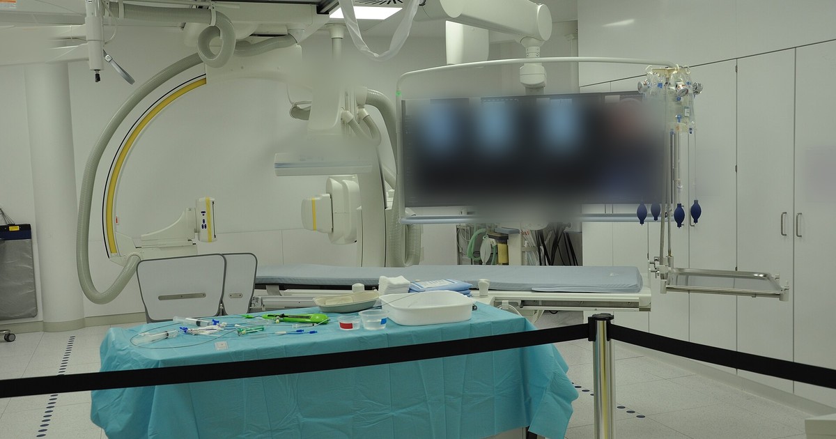 Sala de hemodinâmica vazia com arco em C, monitores suspensos e mesa coberta com campo azul