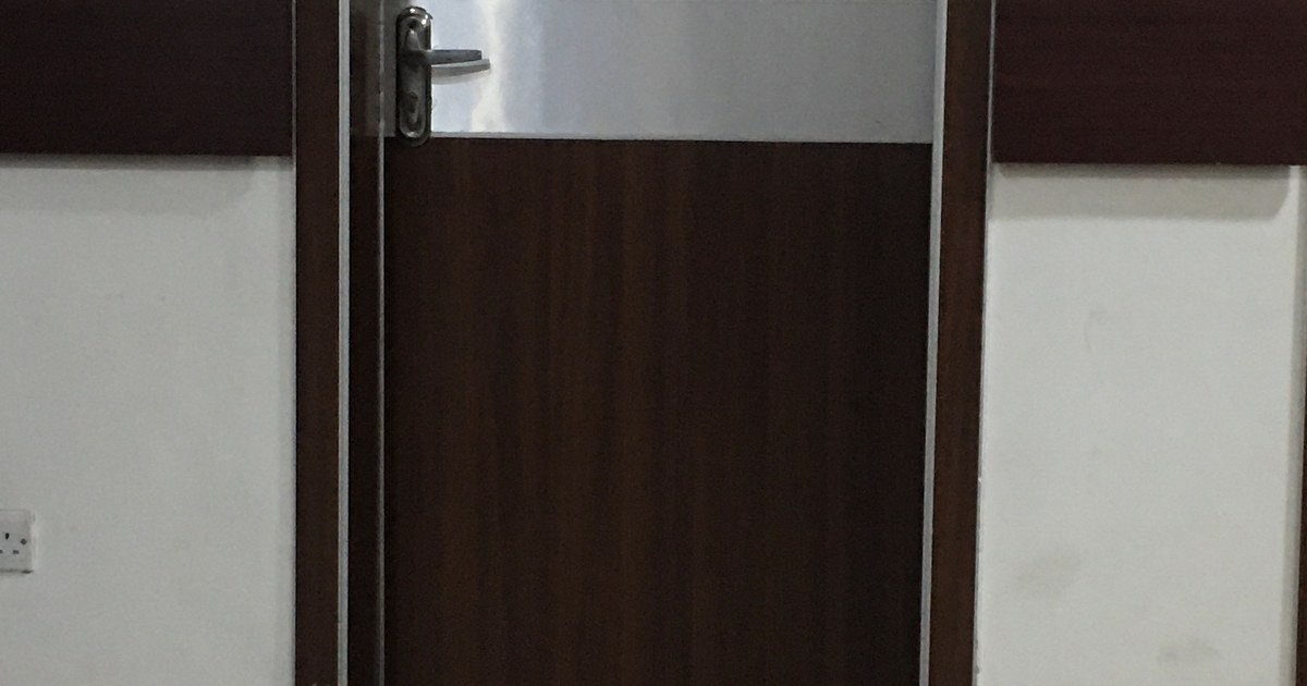 Porta fechada de madeira escura de um consultório de hospital, com maçaneta metálica, entre paredes brancas com faixa marrom