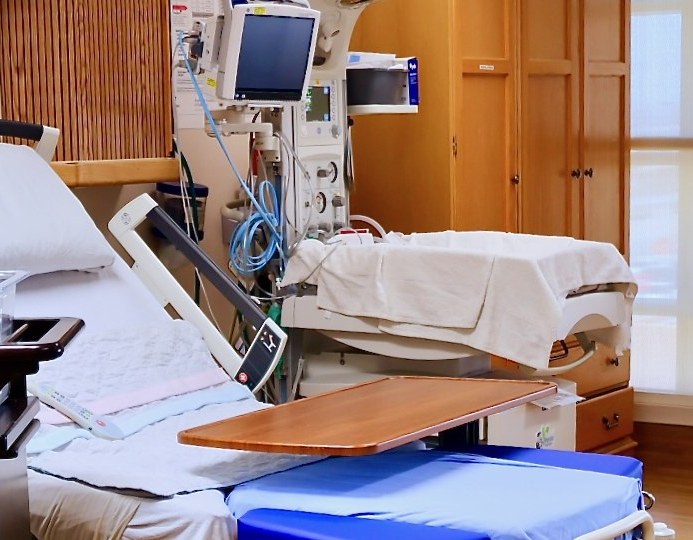 Modernes Krankenhausbett mit über dem Bett stehendem Esstisch, Multiparameter-Monitor und Wandgeräten, im Hintergrund Holzschränke