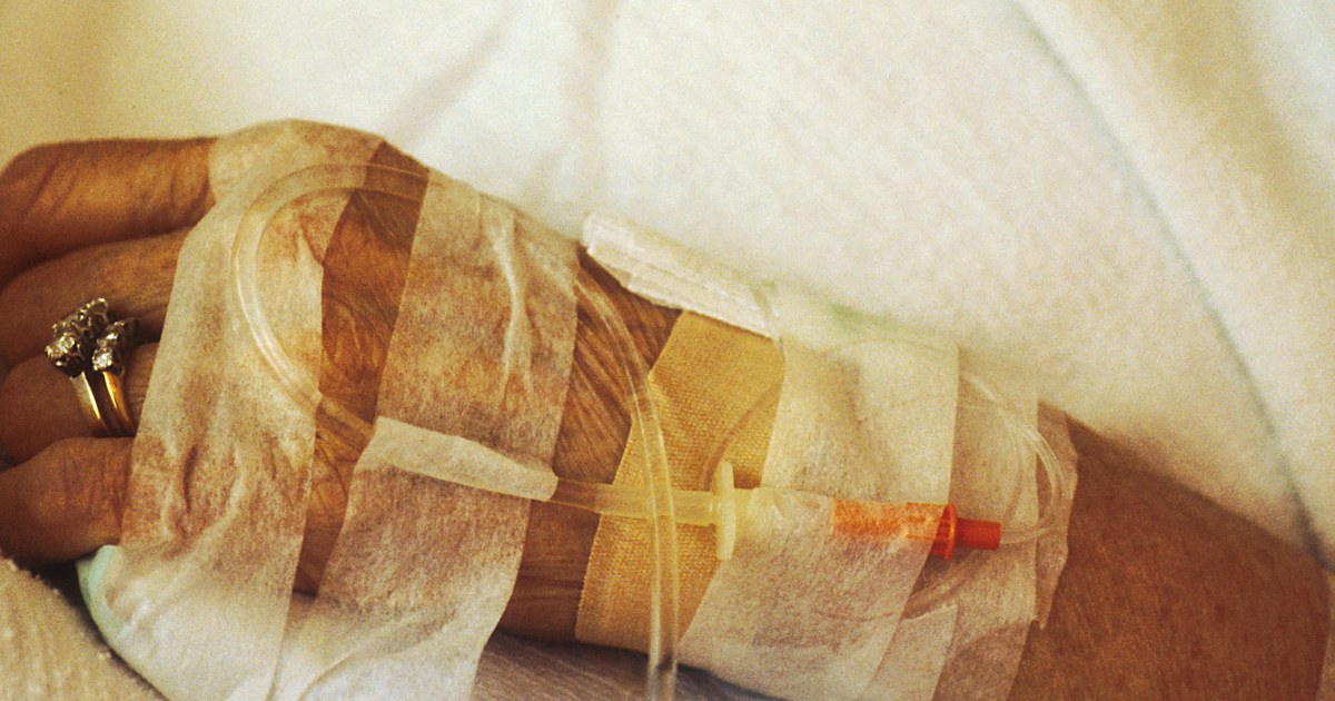 Mão de paciente com acesso venoso fixado por fitas e equipo de infusão sobre o lençol