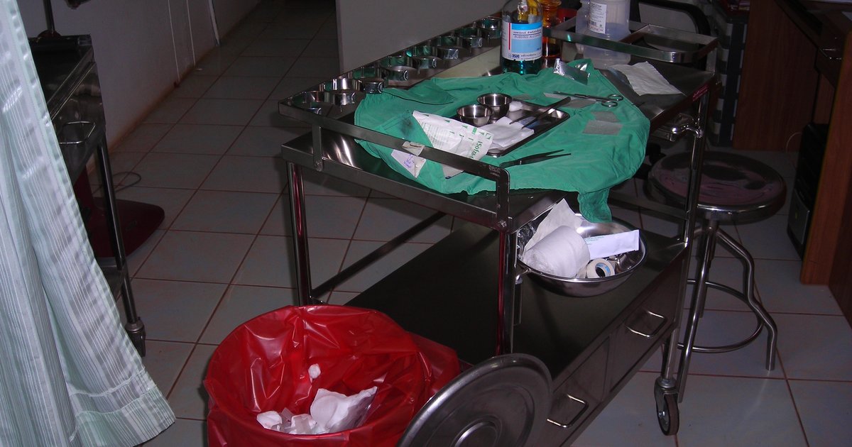 Carro auxiliar de hospital con paño verde, material usado y basurero rojo al lado, cerca de una cortina