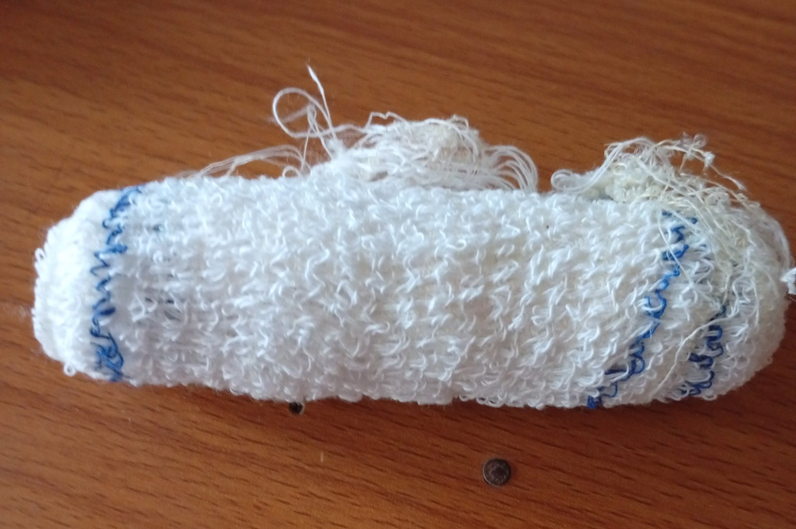 Rolo de atadura de algodão branco, com bordas costuradas em azul e fios soltos, sobre uma mesa de madeira