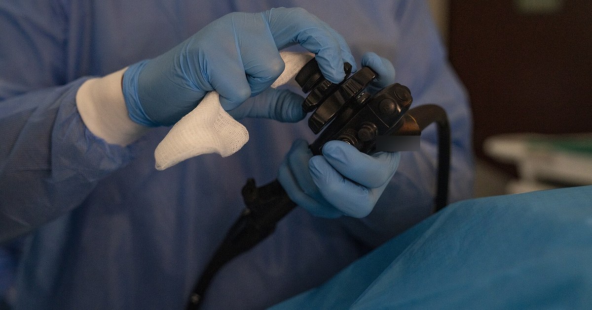 Mãos com luvas azuis seguram o comando de um endoscópio flexível preto durante um exame, com o paciente coberto por campo azul