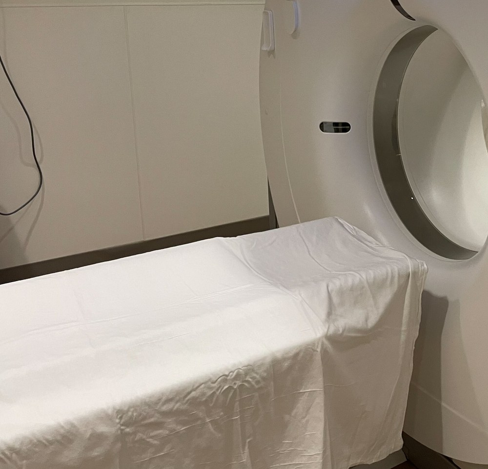 Mesa de exame coberta com lençol branco diante da abertura de um tomógrafo