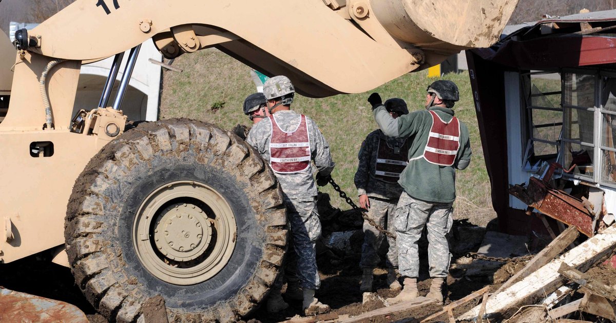Socorristas de capacete e colete revistam escombros de uma casa destruída ao lado de uma grande escavadeira, num exercício de resposta a detonação nuclear