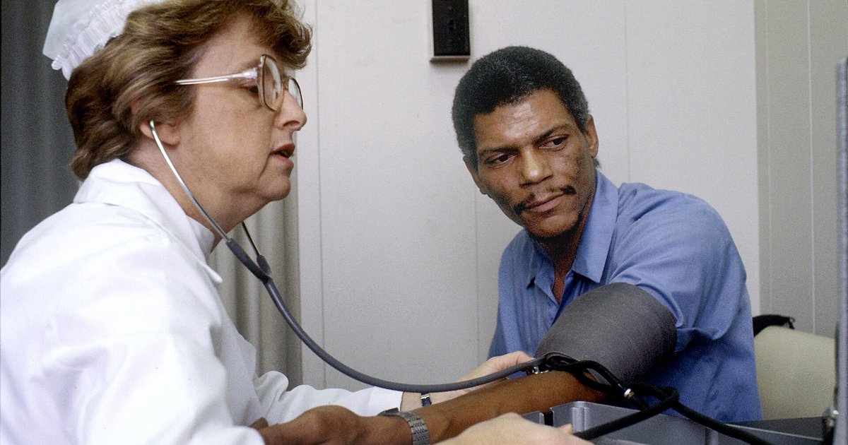 Eine Krankenschwester mit Brille, weißer Haube und weißem Kittel misst mit dem Stethoskop am Ohr den Blutdruck eines Mannes im blauen Hemd; er schaut sie an, den Arm auf dem Tisch abgestützt und die graue Manschette am Arm.