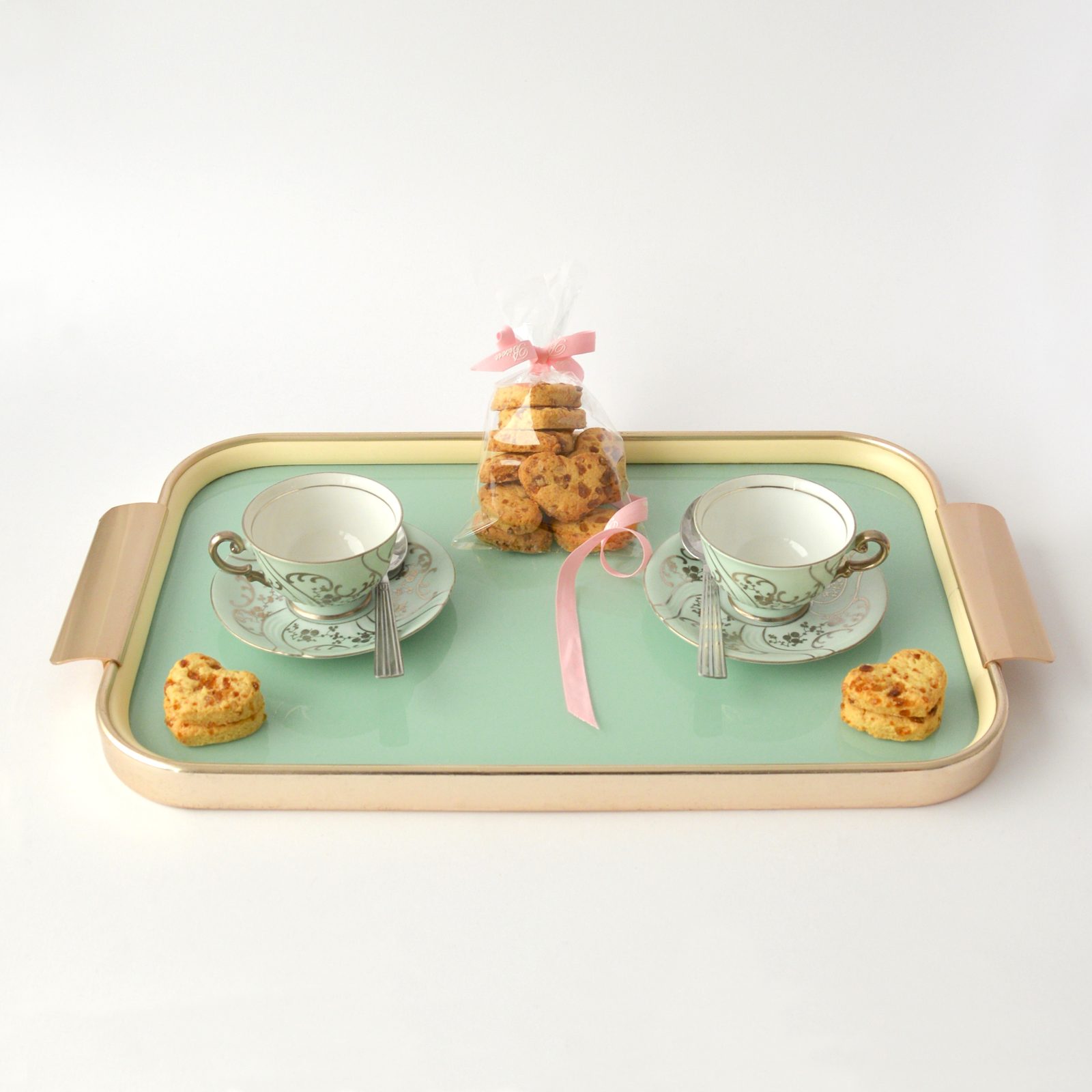 Plateau vert clair avec deux tasses en porcelaine blanche, deux biscuits en forme de cœur et un petit sachet de biscuits avec un nœud rose, sur fond blanc.