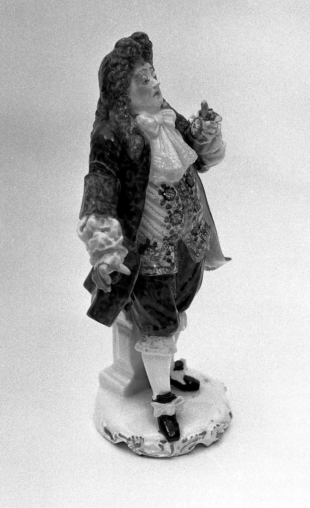 Schwarzweißfoto einer Porzellanfigur eines Arztes aus dem 18. Jahrhundert mit langer gelockter Perücke, dunklem Gehrock, geblümter Weste und Spitzenjabot, der auf einem verzierten Sockel steht und in der erhobenen Hand eine kleine Taschenuhr hält, vor hellem Hintergrund.