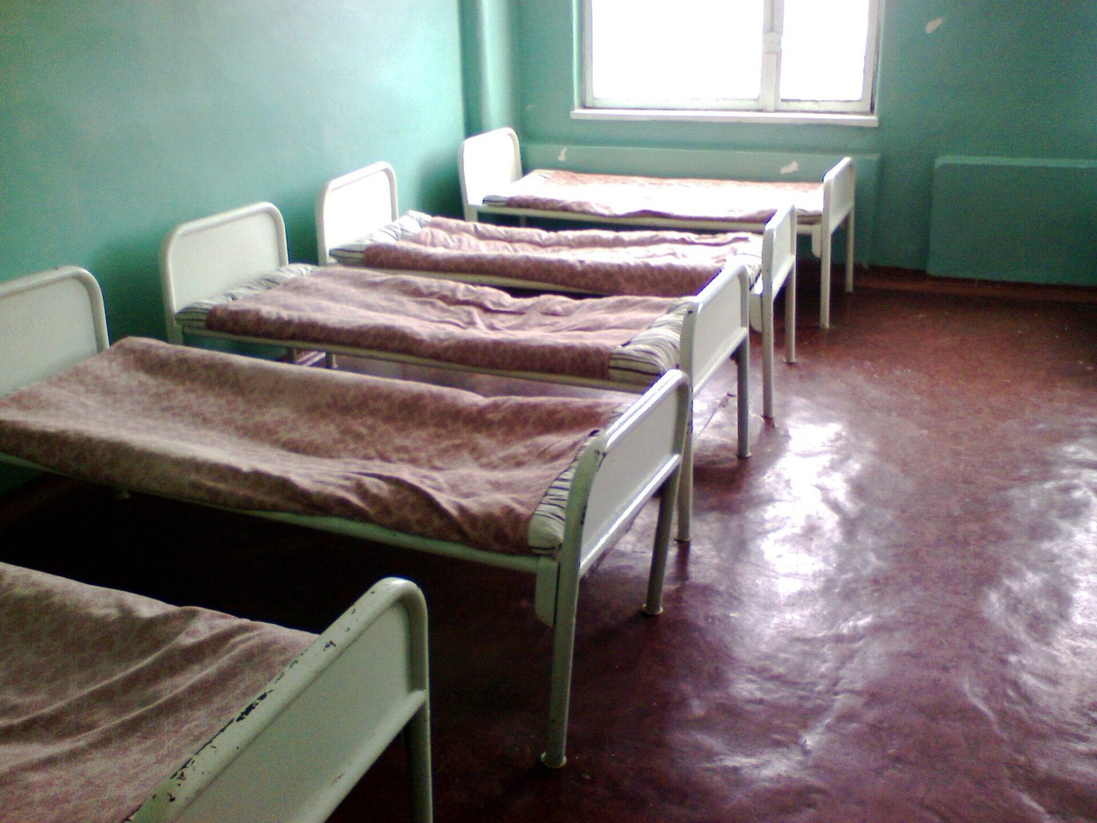 Alter, leerer Krankensaal mit fünf weißen Eisenbetten in einer Reihe, bedeckt mit weinroten Decken, aquagrünen Wänden, rötlichem Boden und einem hellen Fenster im Hintergrund.