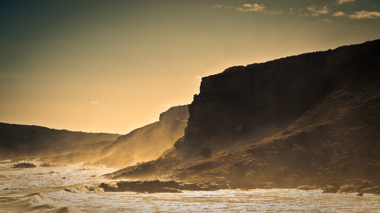 Mar agitado avançando sobre a areia ao entardecer, com um penhasco escuro ao fundo e a luz dourada do sol atravessando a névoa.