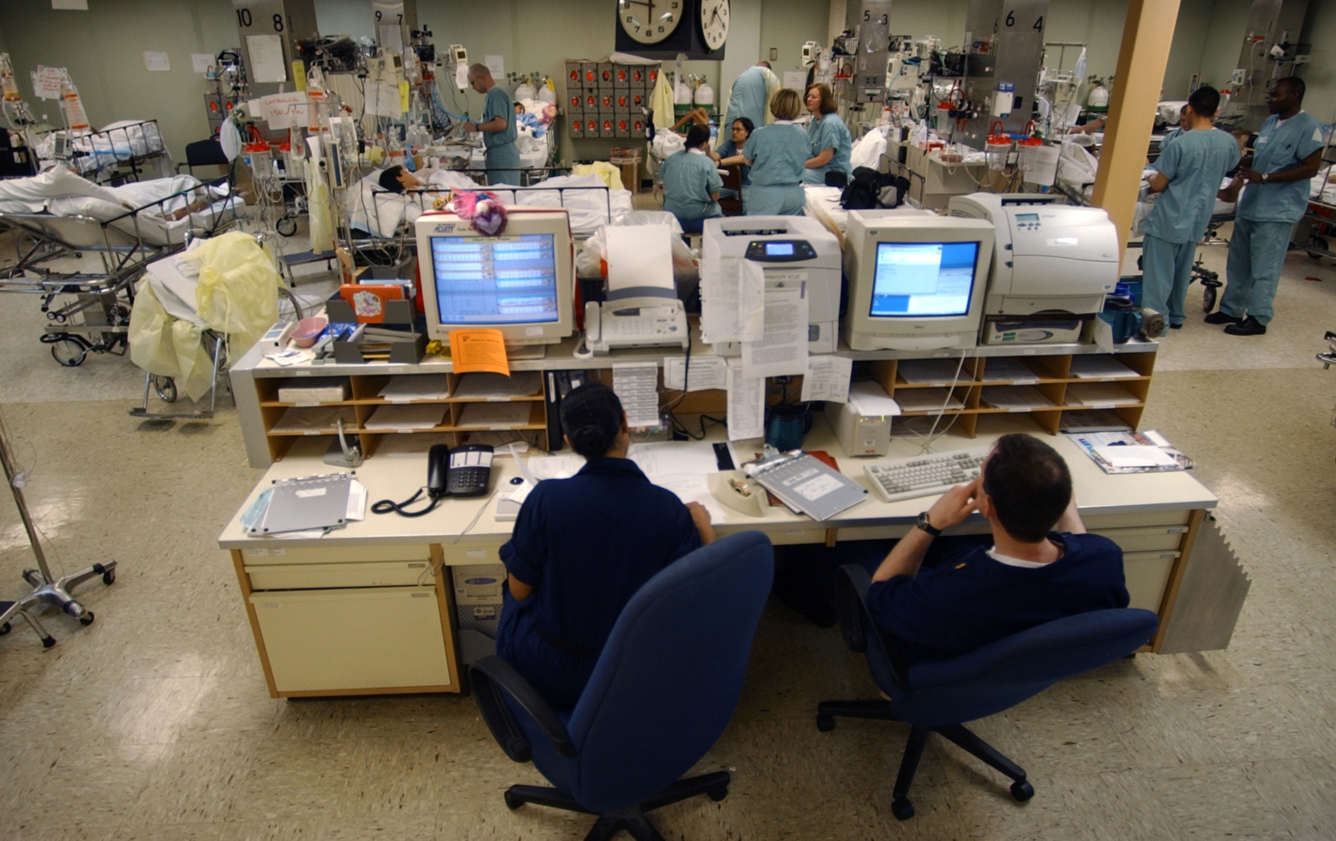Salle de surveillance d'une unité de soins intensifs, avec des professionnels devant des ordinateurs et des lits au fond.