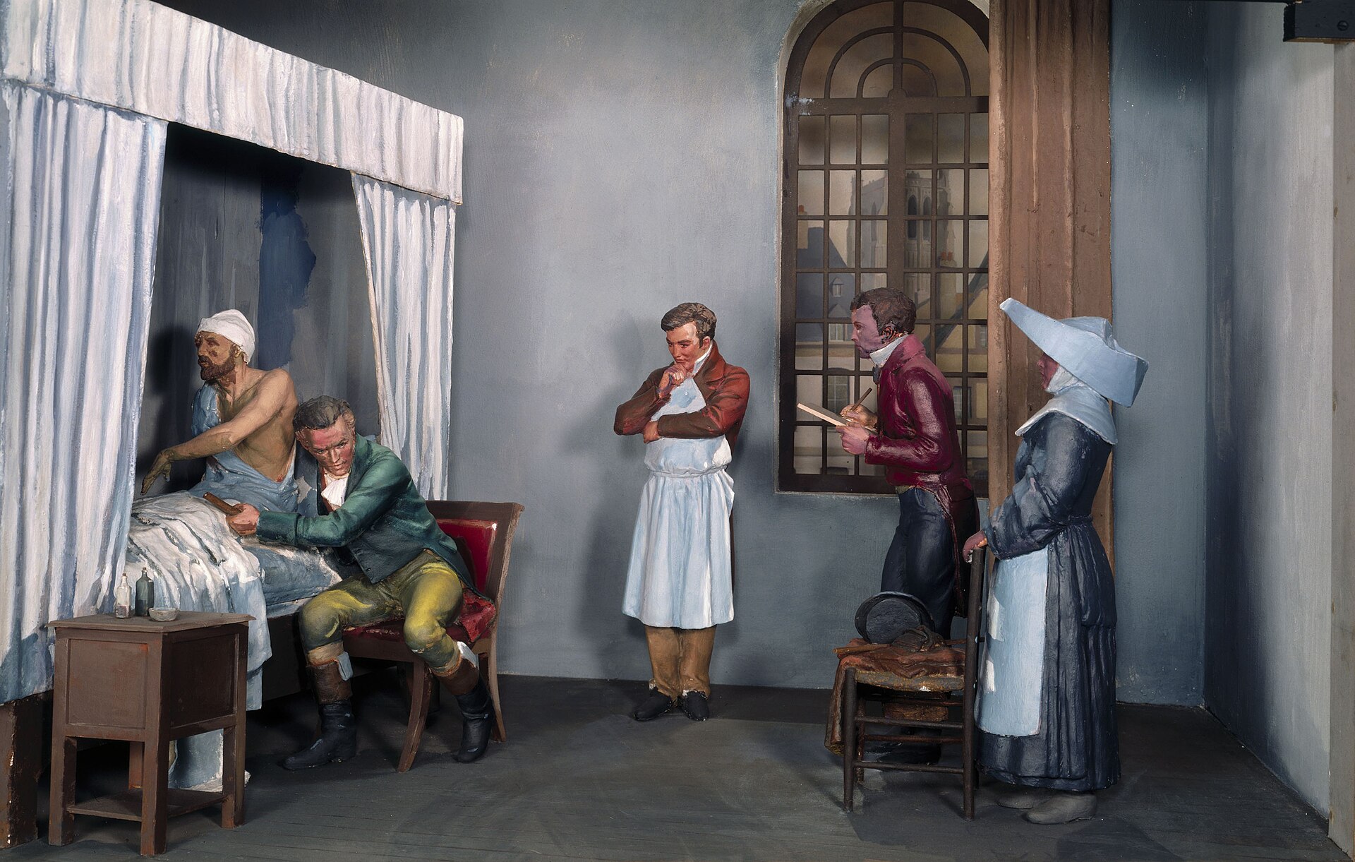 Ancien diorama d'une salle d'hôpital avec un médecin assis qui appuie sa tête contre la poitrine d'un patient alité, tandis que des collègues et une religieuse observent.