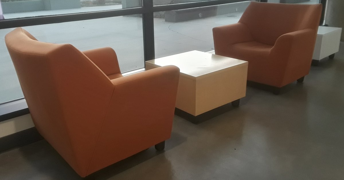 Duas poltronas laranja vazias e uma mesa baixa clara diante de uma janela ampla, num corredor iluminado.