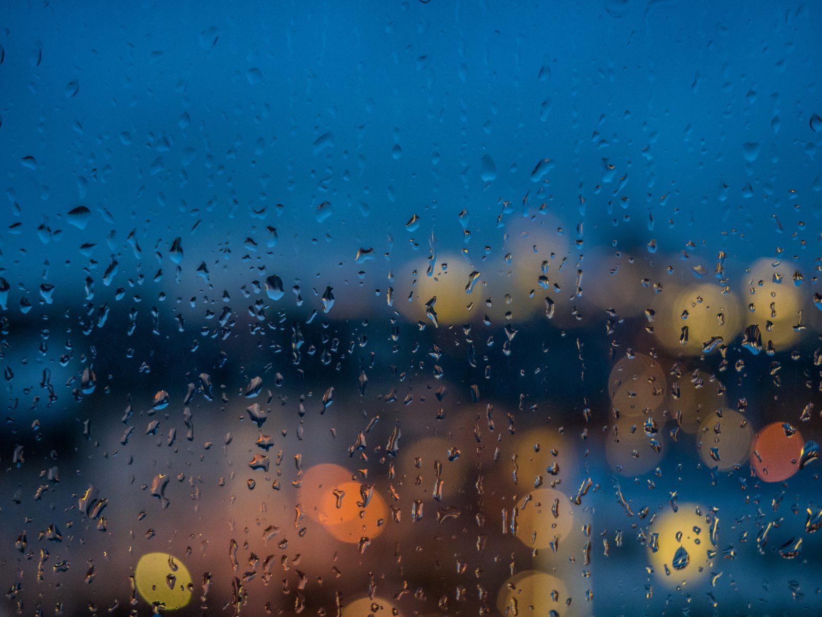 Gotas de chuva numa vidraça, com luzes desfocadas da rua ao fundo num fim de tarde azulado.