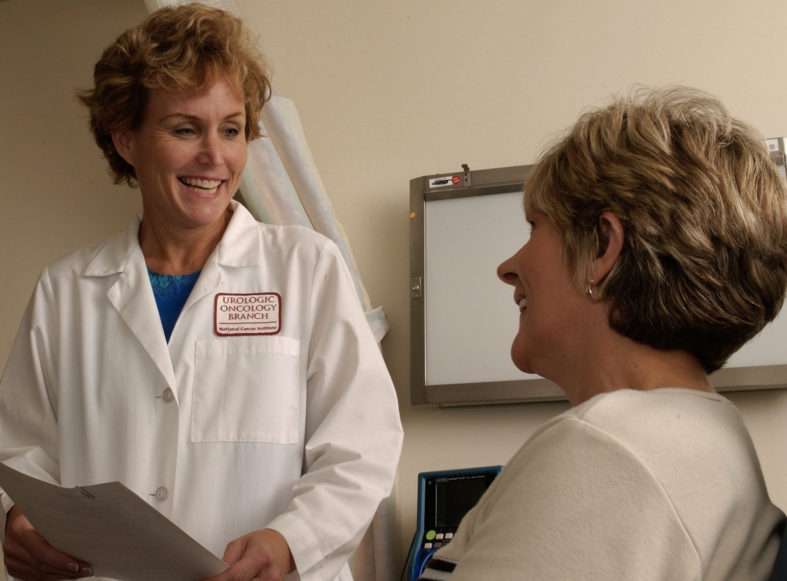 A doctor in a white coat smiles while holding papers as she talks to a short-haired patient, seen in profile and also smiling, in an office with a light box on the wall.