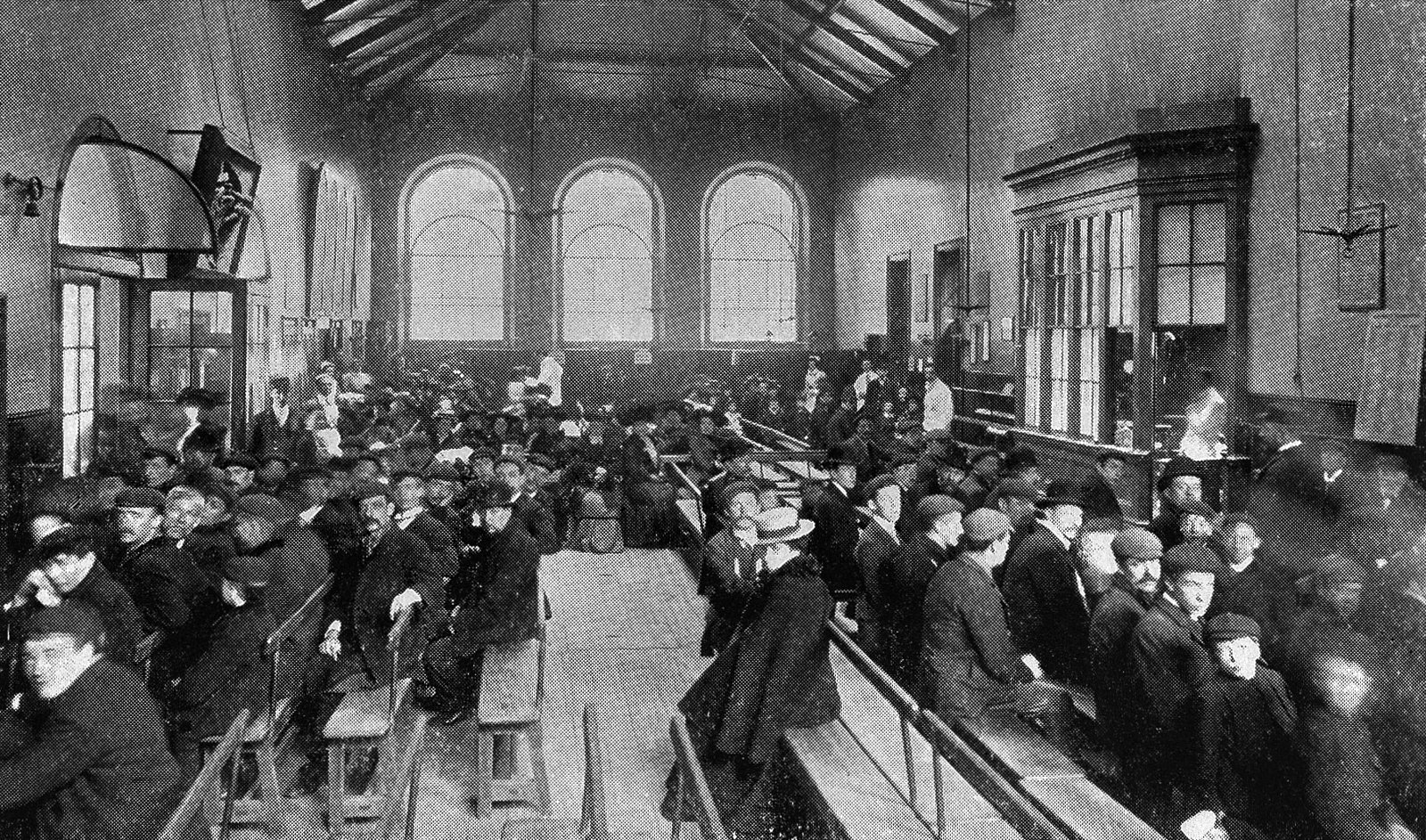 An old black-and-white photo of a crowded waiting room in a London hospital: dozens of men and women in hats and dark coats sit on long wooden benches, under a high ceiling with iron trusses and arched windows in the background.