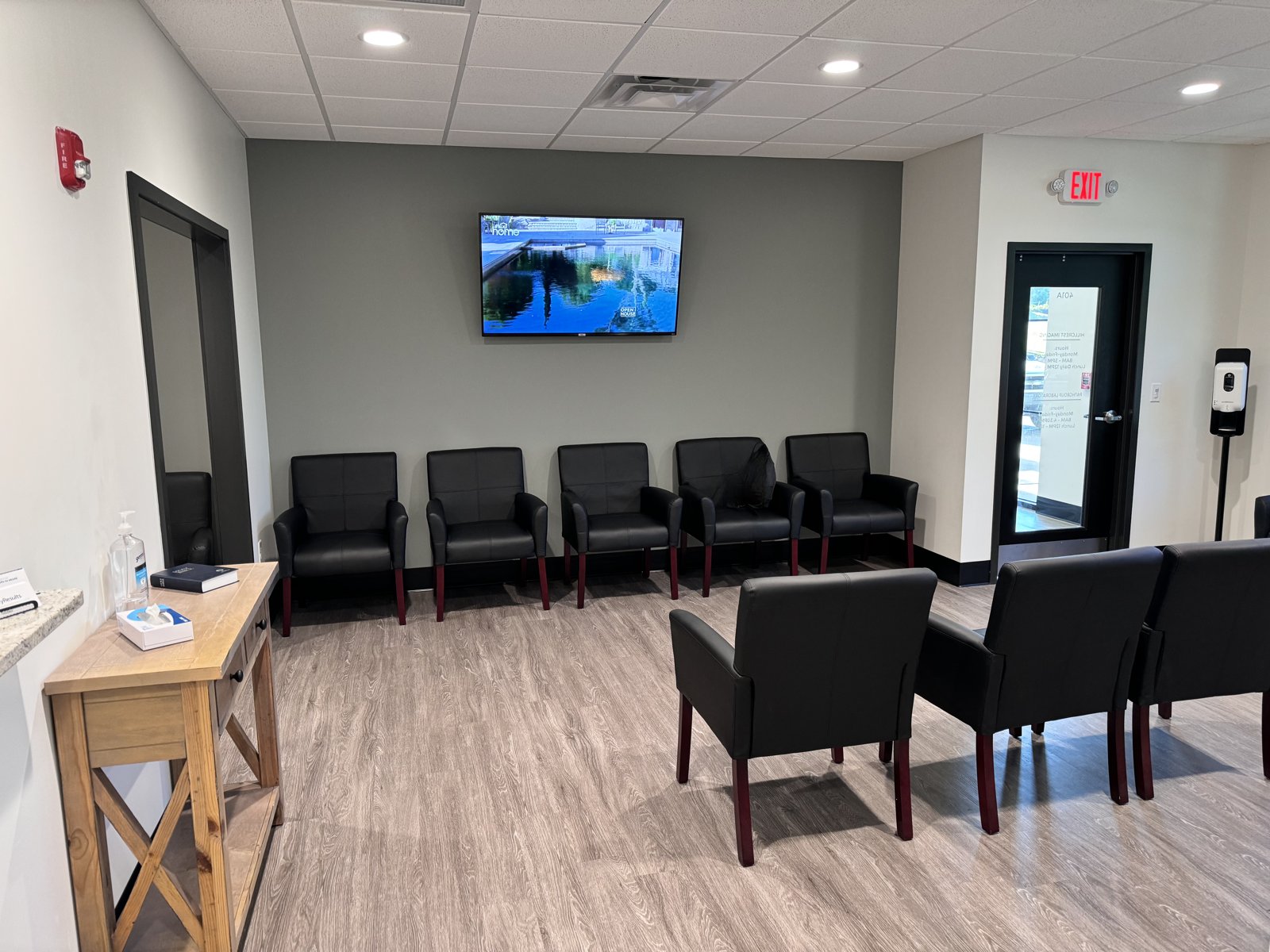 A clinic waiting room with black leather chairs against gray walls, a wooden table with leaflets, a television on the wall and an exit door in the background.