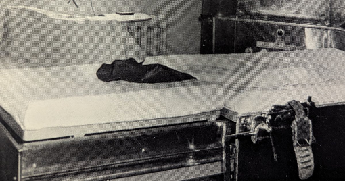 Fotografia antiga em preto e branco de uma mesa de parto vazia, coberta com lençol branco, em uma sala de hospital