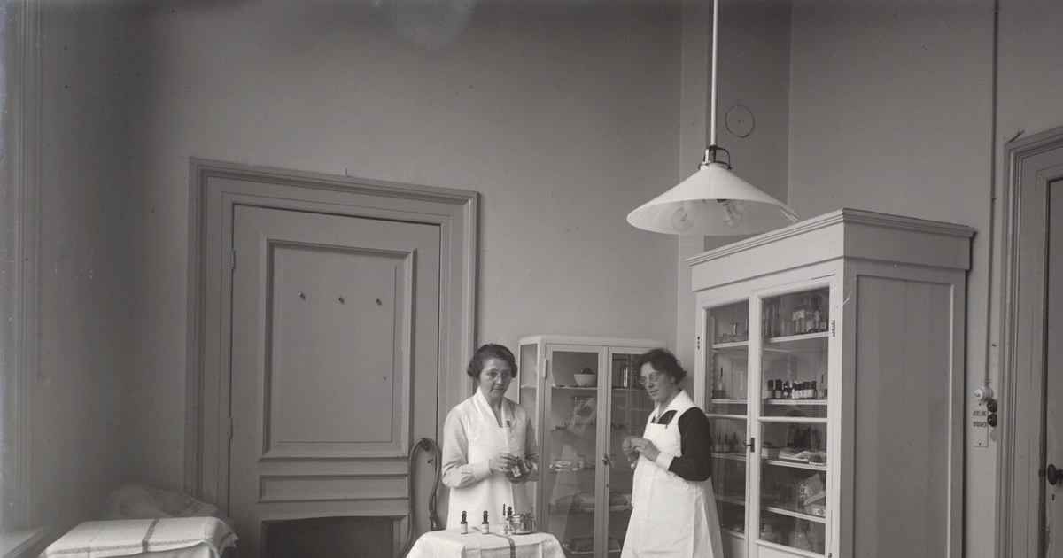 Fotografía antigua en blanco y negro de una sala de tratamiento hospitalario con una camilla cubierta por sábanas a rayas en el centro, dos mujeres con delantal blanco de pie junto a un armario de vidrio con frascos, una lámpara de araña colgante y puertas de madera.