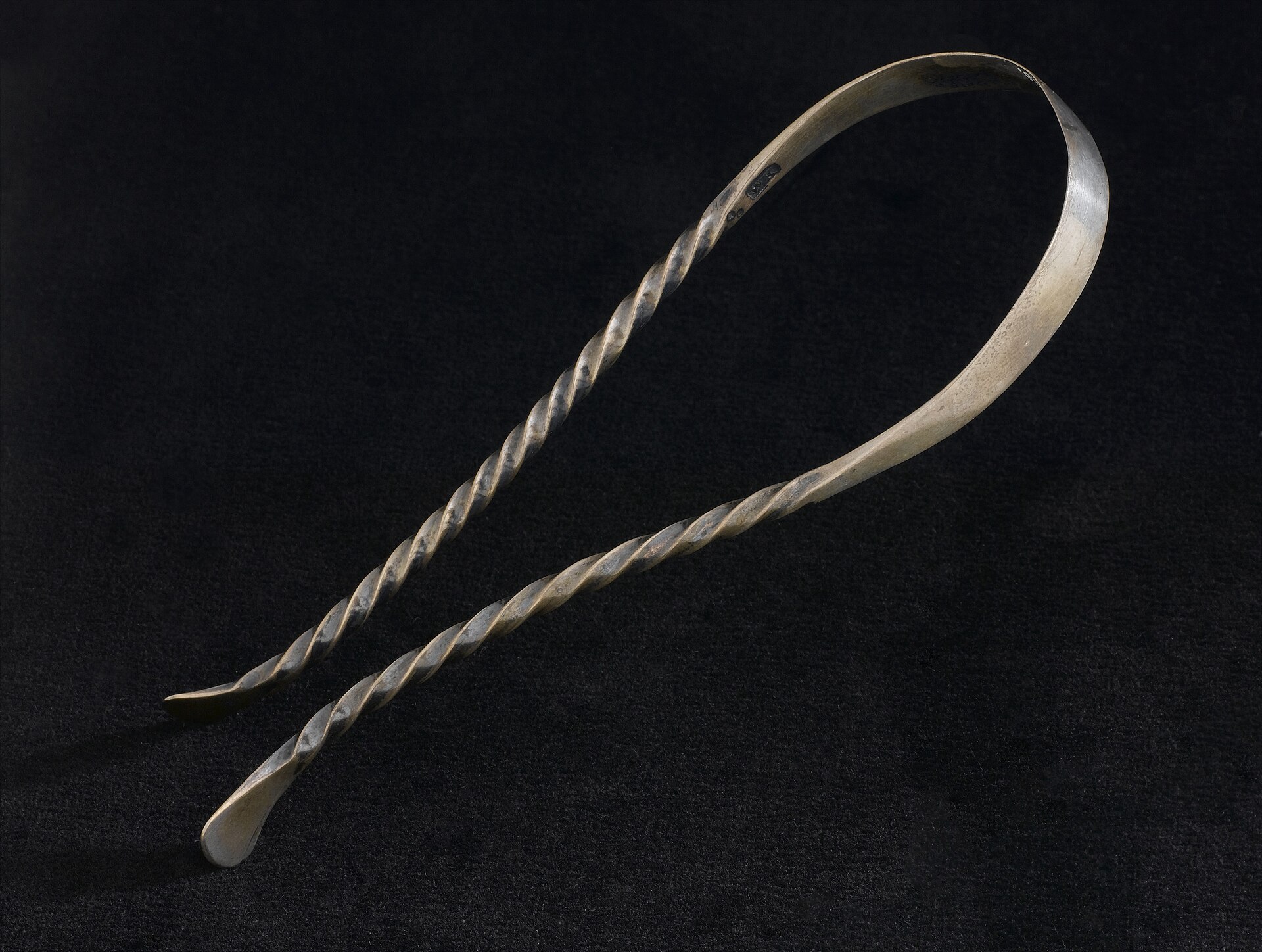 Ancien grattoir à langue en argent, une fine bande pliée en arc dont les deux tiges sont torsadées en spirale, posé sur fond noir.