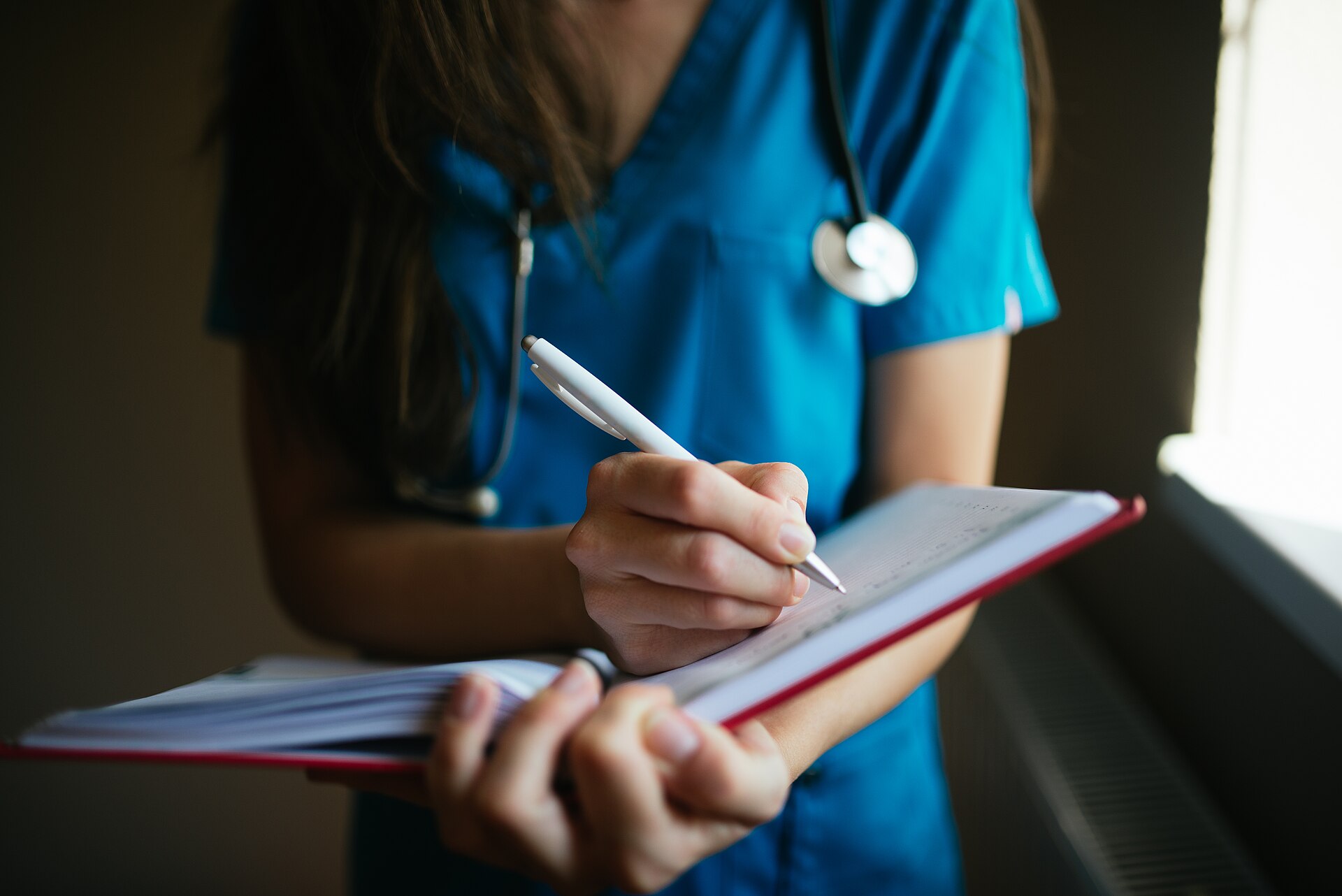 Professionnel de santé en uniforme bleu, stéthoscope autour du cou, écrivant au stylo blanc dans un carnet à couverture rouge qu'il tient ouvert de l'autre main, près d'une fenêtre éclairée.