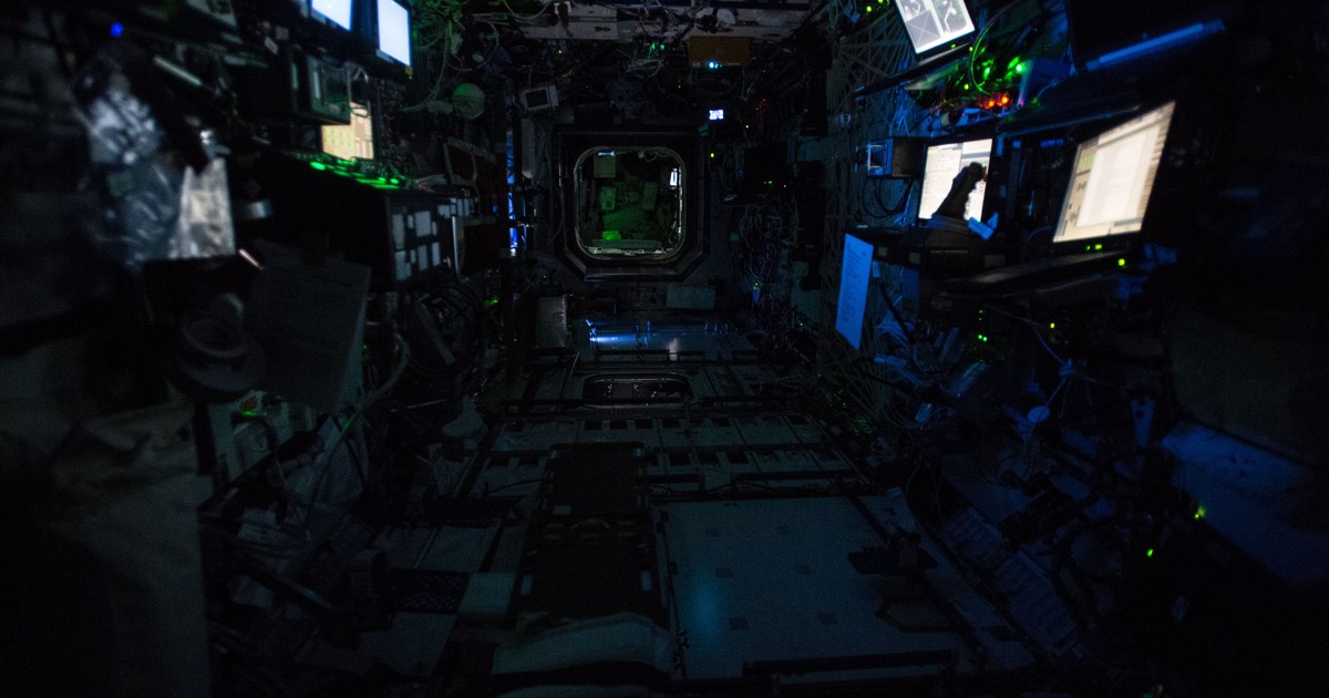 Interior do laboratório Destiny da Estação Espacial Internacional na penumbra, com equipamentos, cabos e telas acesas