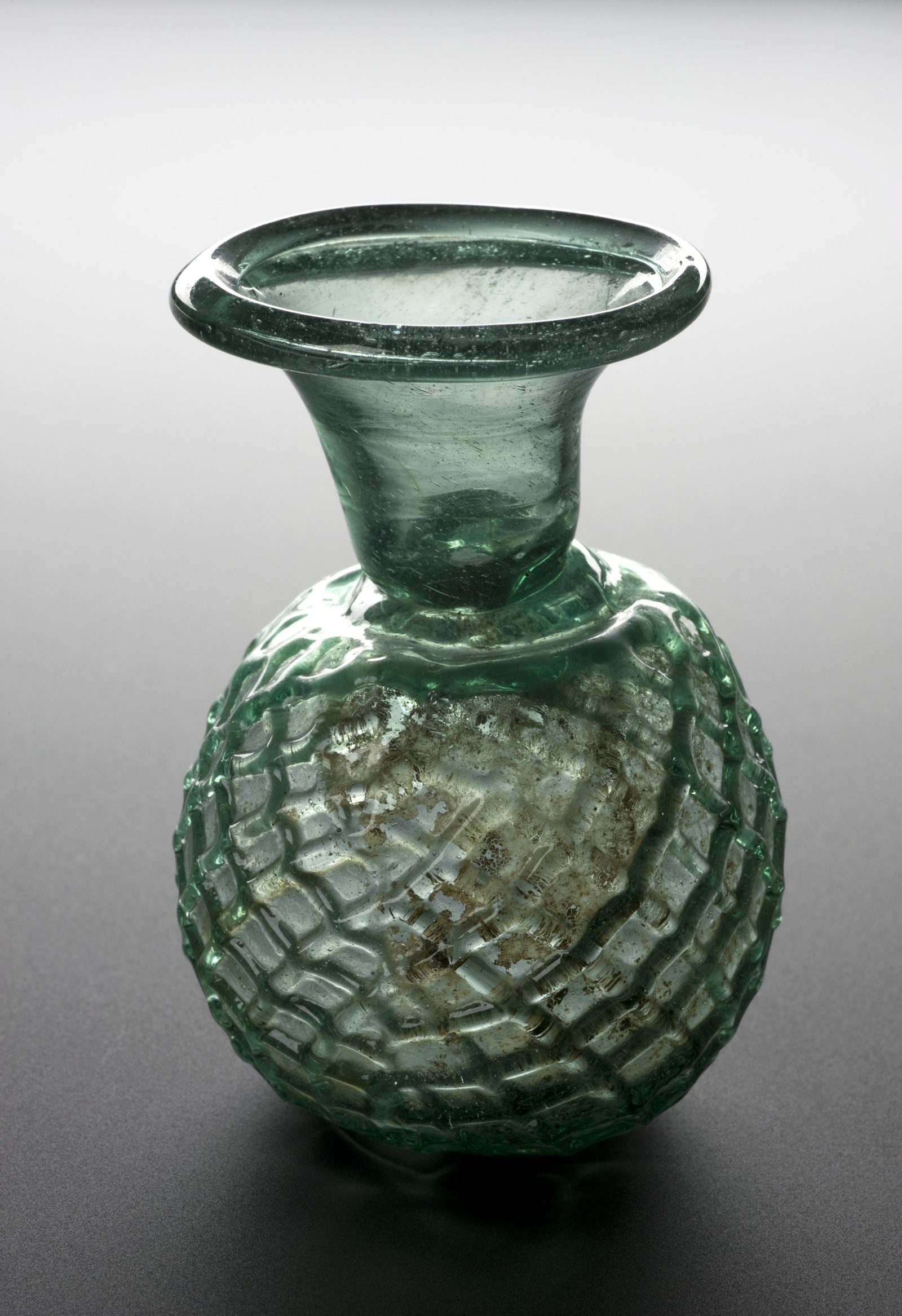 Small Roman green glass flask, with a wide, thick-rimmed neck and a round body covered by a net-shaped relief, on a gray background.