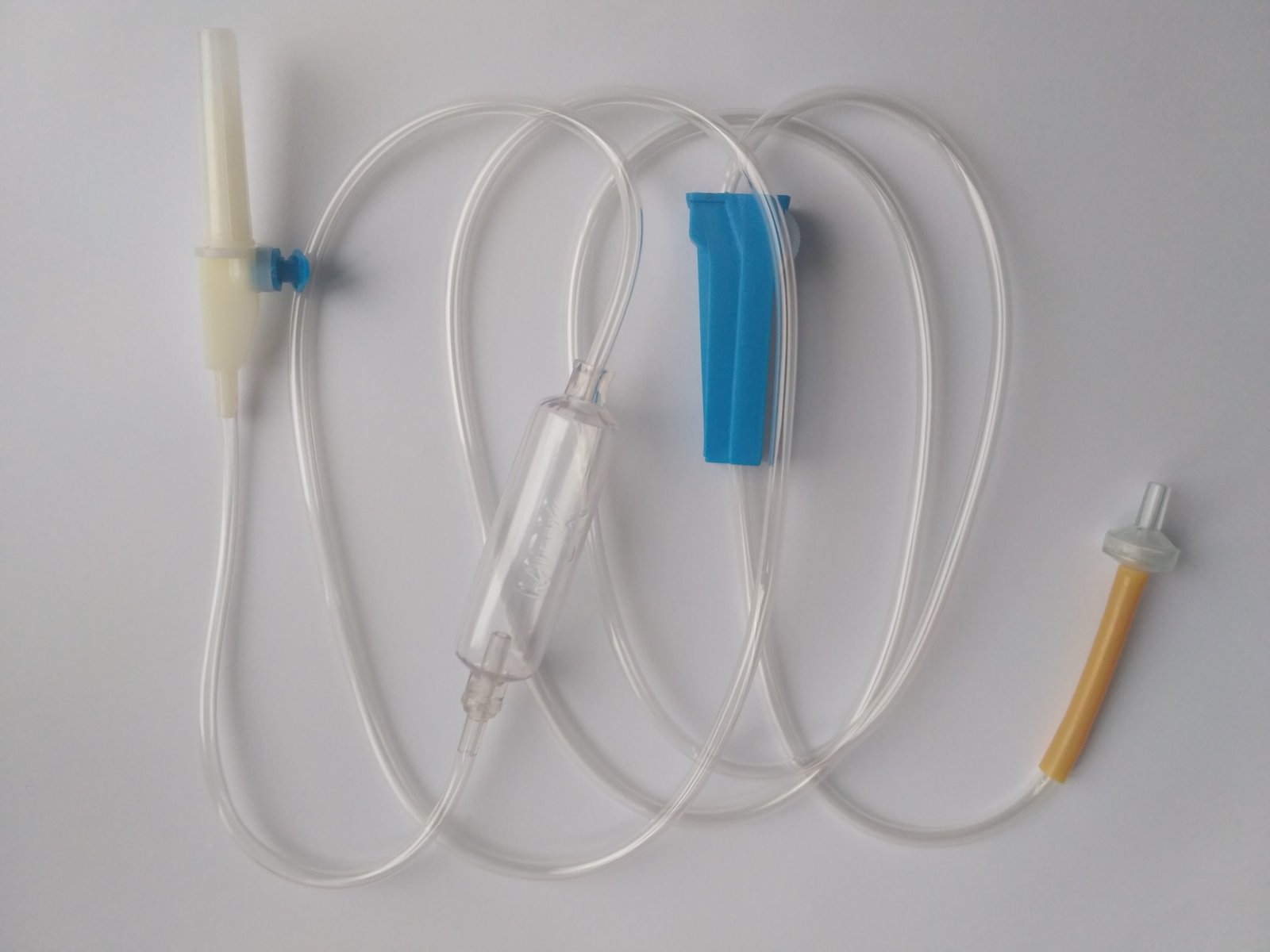 Disposable IV infusion set laid out on a white background, with the piercing spike, the clear drip chamber, the blue regulating clamp and the yellow connector at the end.