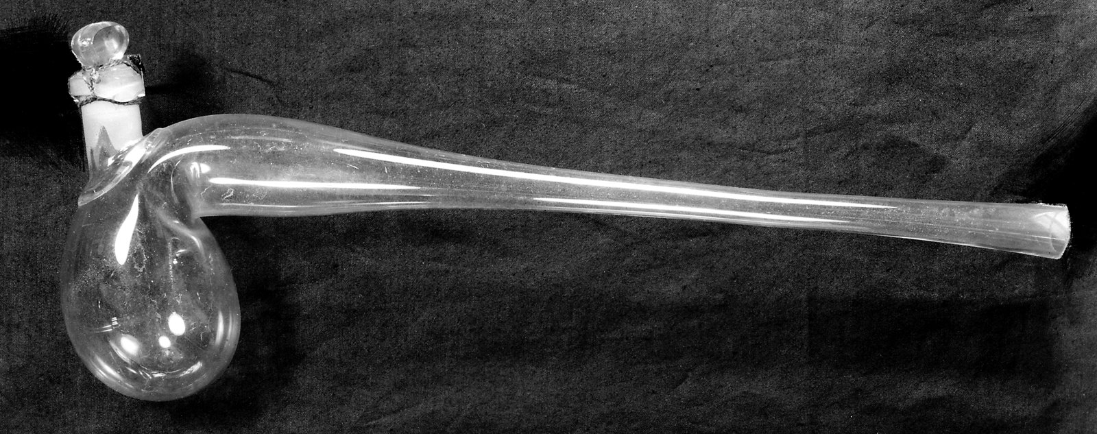 19th-century clear glass retort, with the rounded bulb and glass stopper on the left and a long tapered spout extending to the right, on dark fabric.