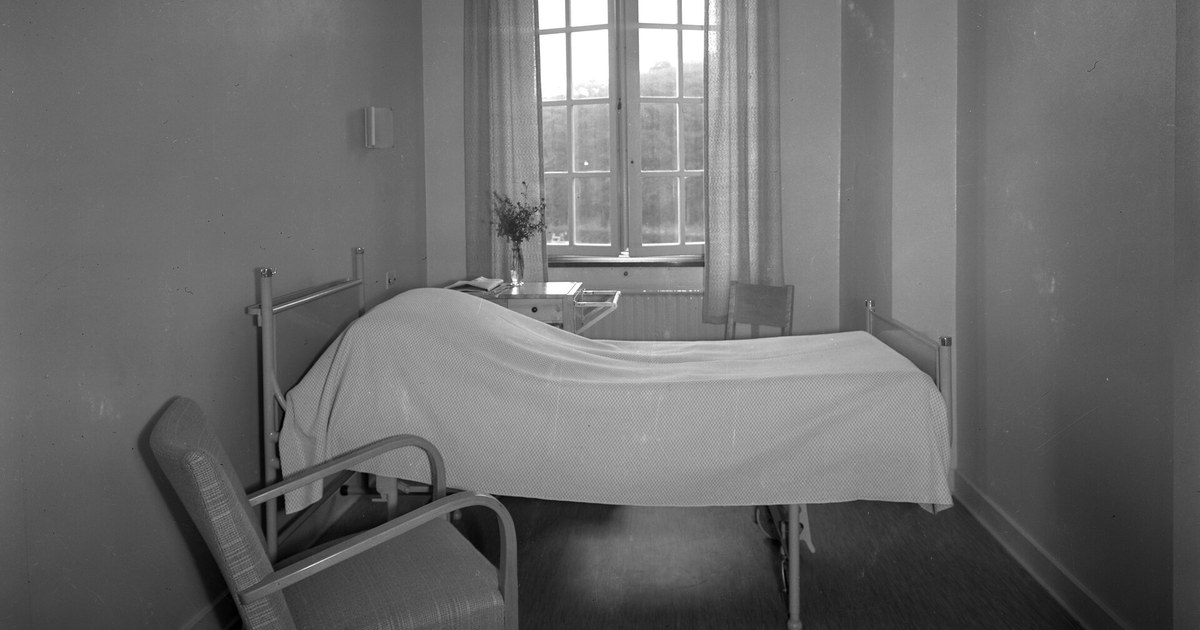 Quarto de hospital antigo em preto e branco com leito arrumado, cadeira e janela ao fundo