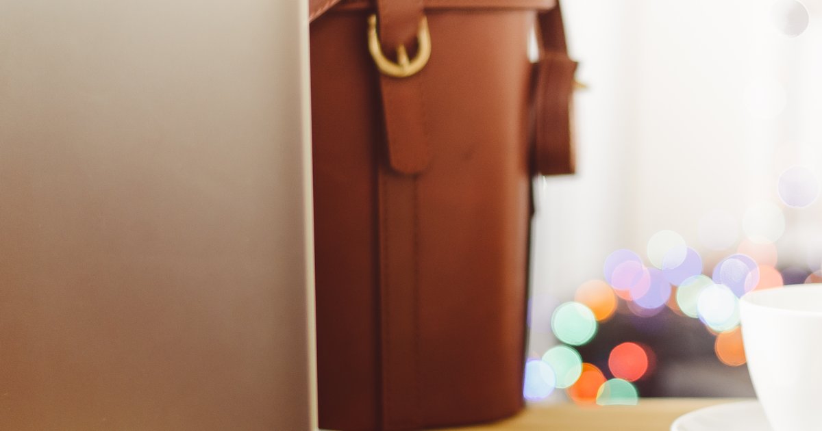 Bolsa de couro marrom ao lado de um notebook e de uma xícara branca sobre mesa de madeira com luzes desfocadas ao fundo