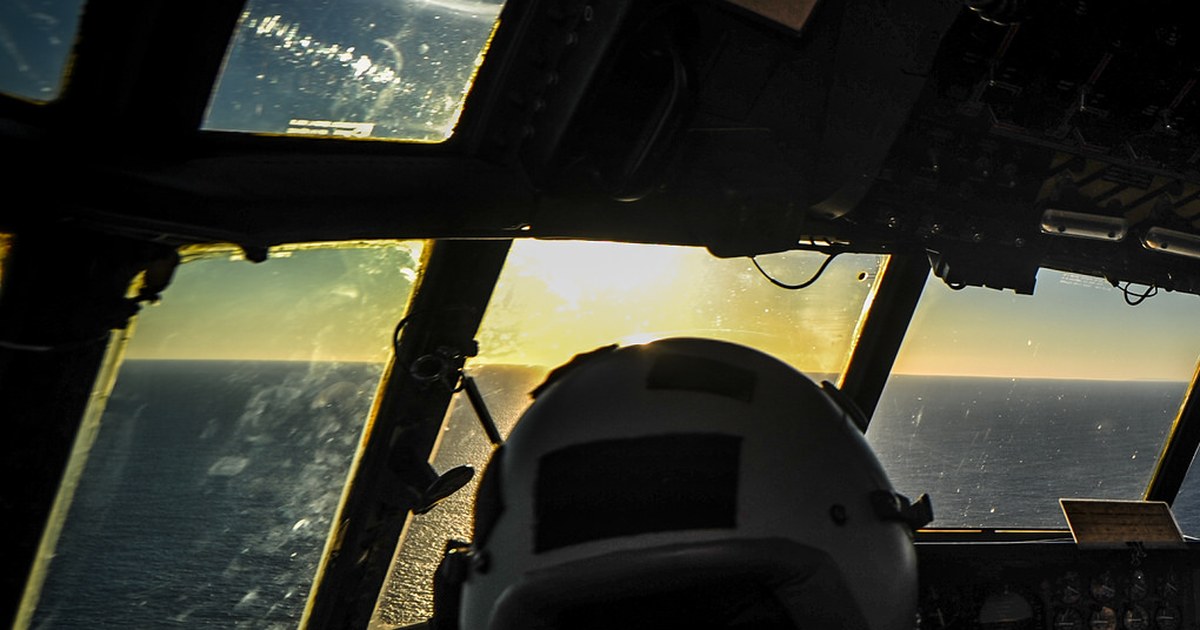 Cabine de avião em voo sobre o mar ao entardecer, vista por trás do capacete de um tripulante