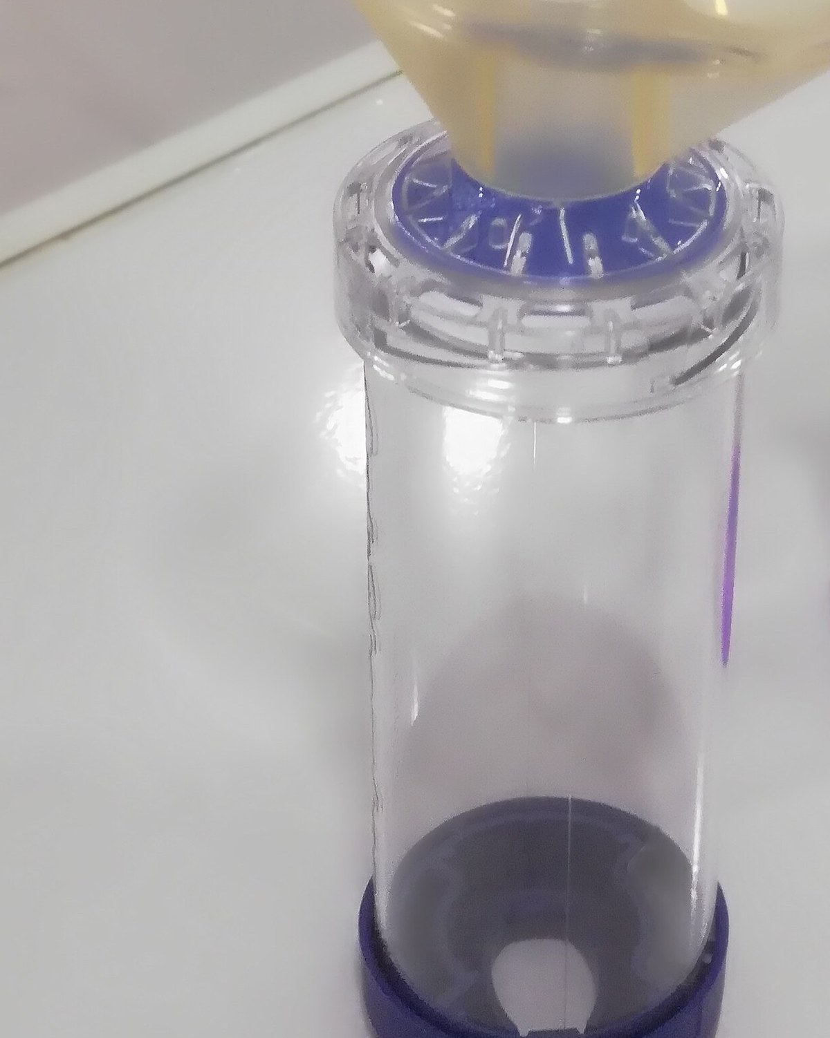 Espaçador transparente com máscara e válvula azul, usado para inalação, sobre bancada branca
