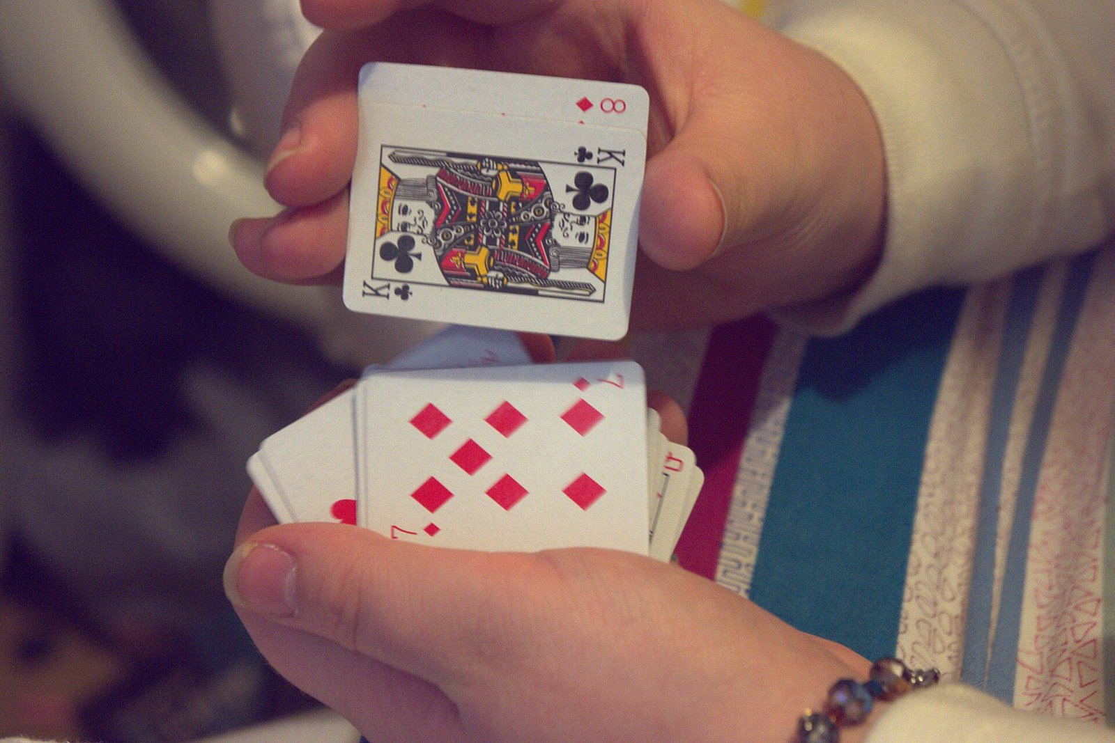Hände, die kleine Spielkarten mischen: in der oberen Hand ein Kreuz-König, dahinter eine Karo-Acht; in der unteren Hand Karo-Karten.