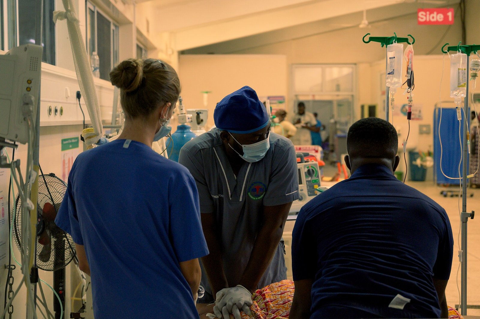 In a hospital emergency room, a professional wearing a blue cap, mask and gloves performs chest compressions on a patient covered by a patterned cloth, while a colleague in blue clothing and a mask and a man in a navy blue shirt watch closely; around them, stands with IV bags, monitors and a sign reading Side 1.