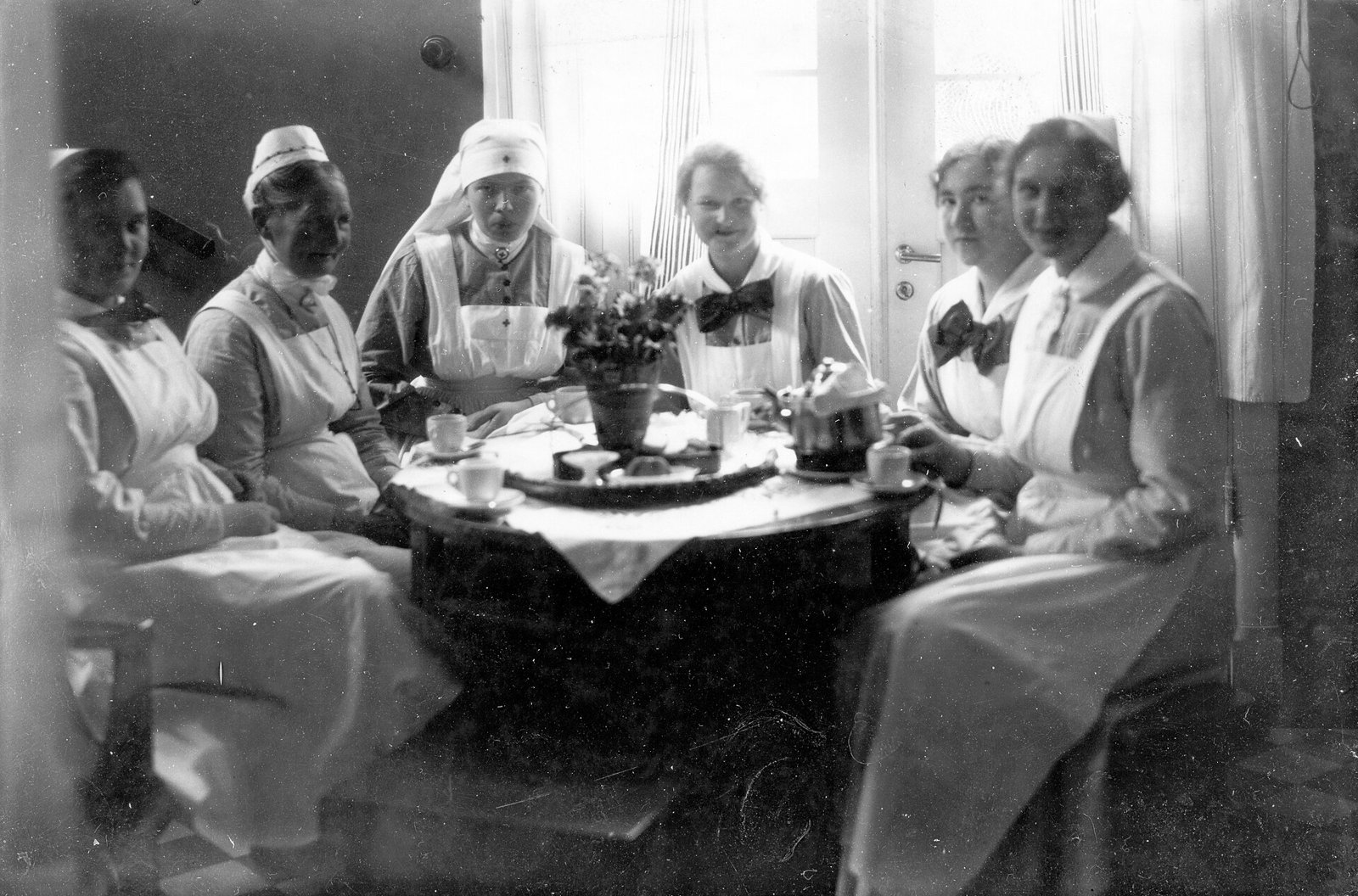 Old black and white photo of six nurses and nursing students, in white aprons, collars with bows and caps, sitting around a round table with cups, a teapot and a vase of flowers; some smile at the camera, in front of a window with light curtains.