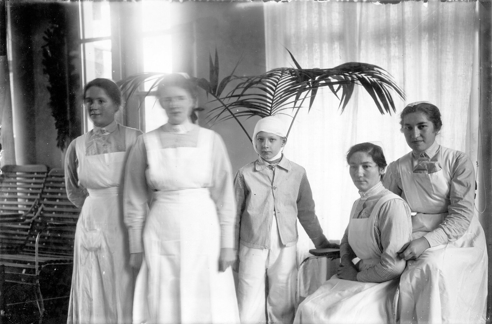 Old black and white photo of four nurses in white aprons and caps gathered in a hospital room with a palm tree and light curtains: two are standing on the left, two are sitting on the right, and between them a boy in striped pajamas with his head bandaged.