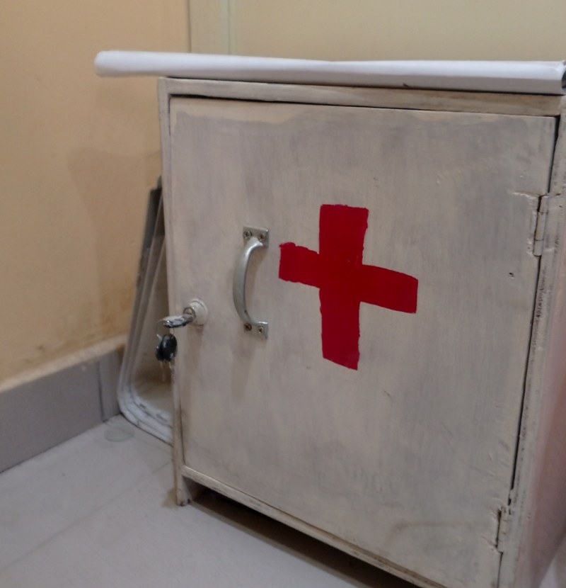 Armário metálico de primeiros socorros com uma cruz vermelha pintada na porta, num canto de posto de saúde