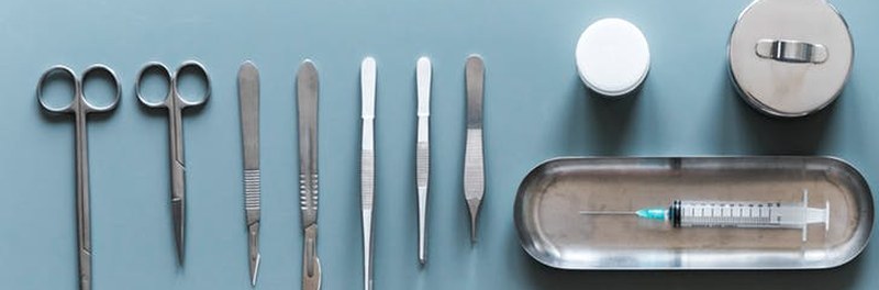 Tesouras, pinças e bisturis enfileirados ao lado de uma cuba metálica com uma seringa