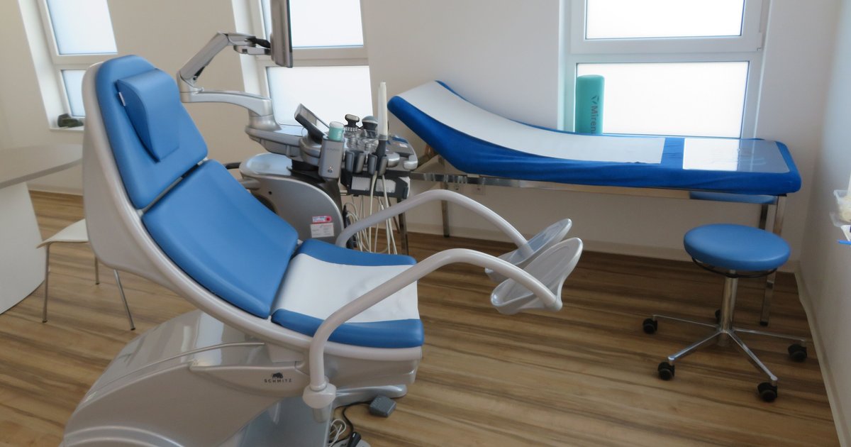 Consultório ginecológico vazio com cadeira de exame azul e branca, maca azul e banqueta junto à janela