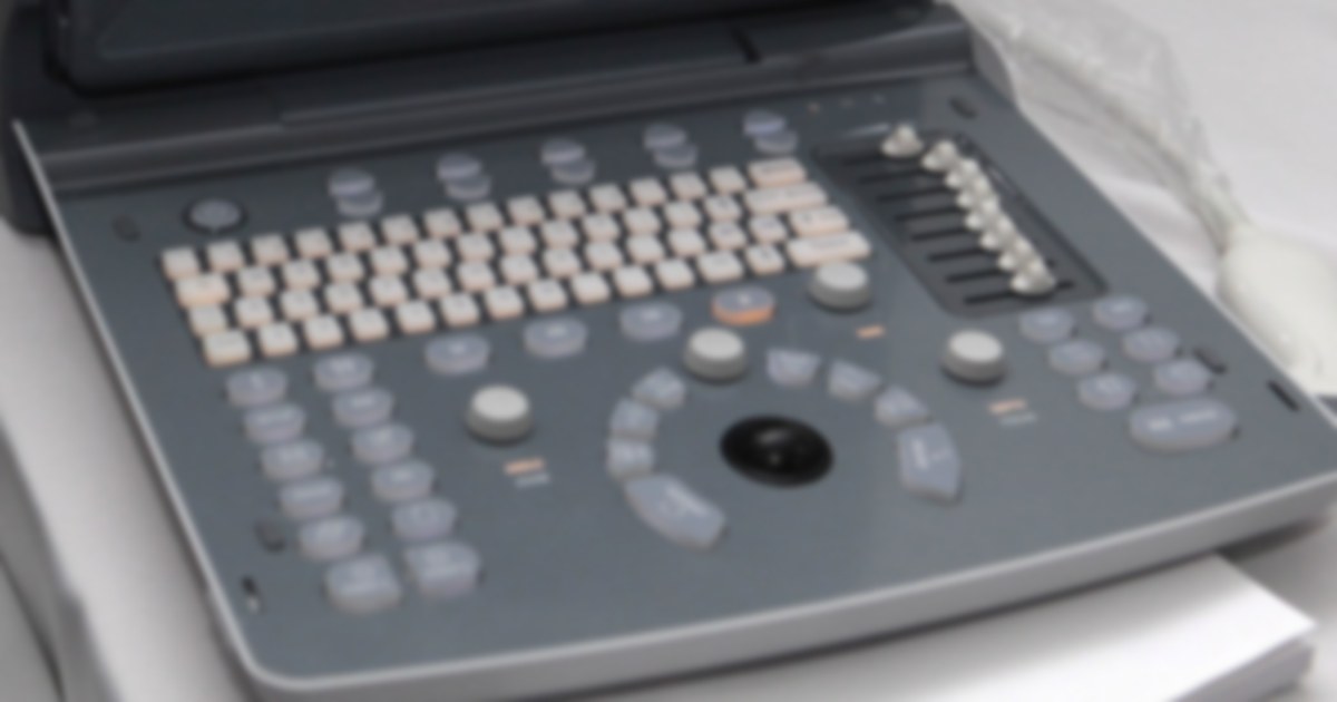 Painel de controle de um aparelho de ultrassom, com teclado, botões de ajuste e trackball, e o transdutor apoiado ao lado