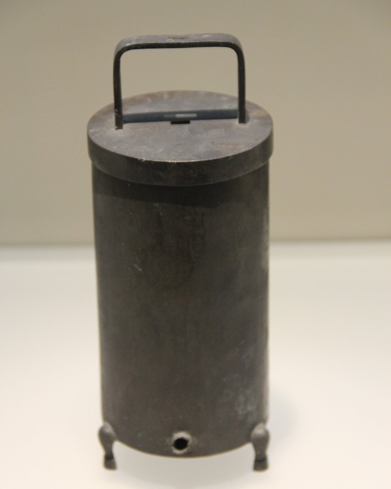 Recipiente cilíndrico escuro de metal com tampa, alça e três pés curtos, com um pequeno orifício perto da base, exposto sobre fundo claro