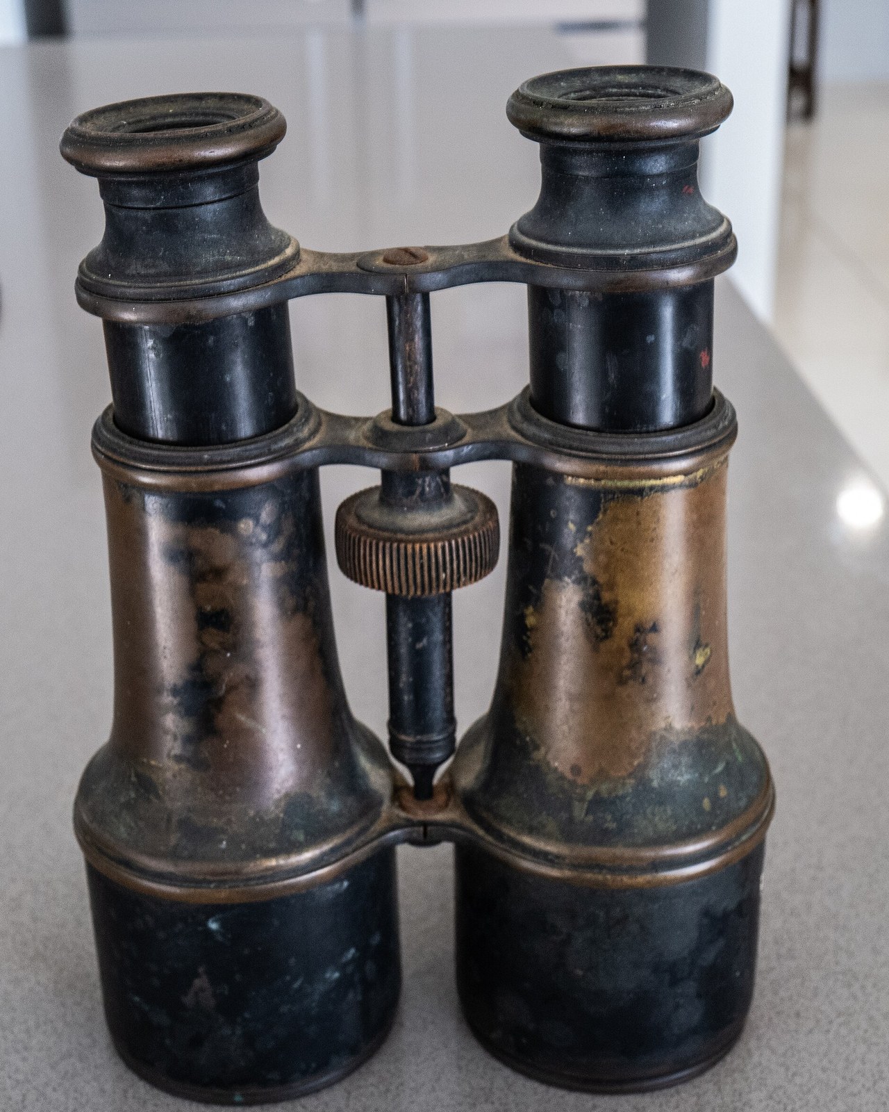 Binóculo antigo de metal escuro com partes de latão gastas e roda central de foco, em pé sobre uma bancada cinza