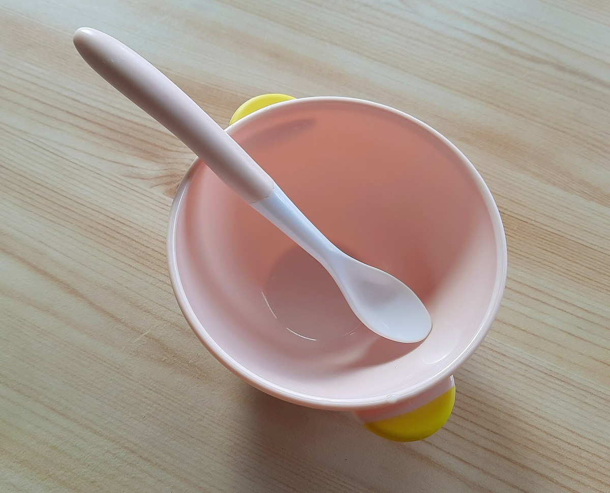 Tigela plástica rosa com uma colher branca apoiada dentro, sobre uma mesa de madeira clara